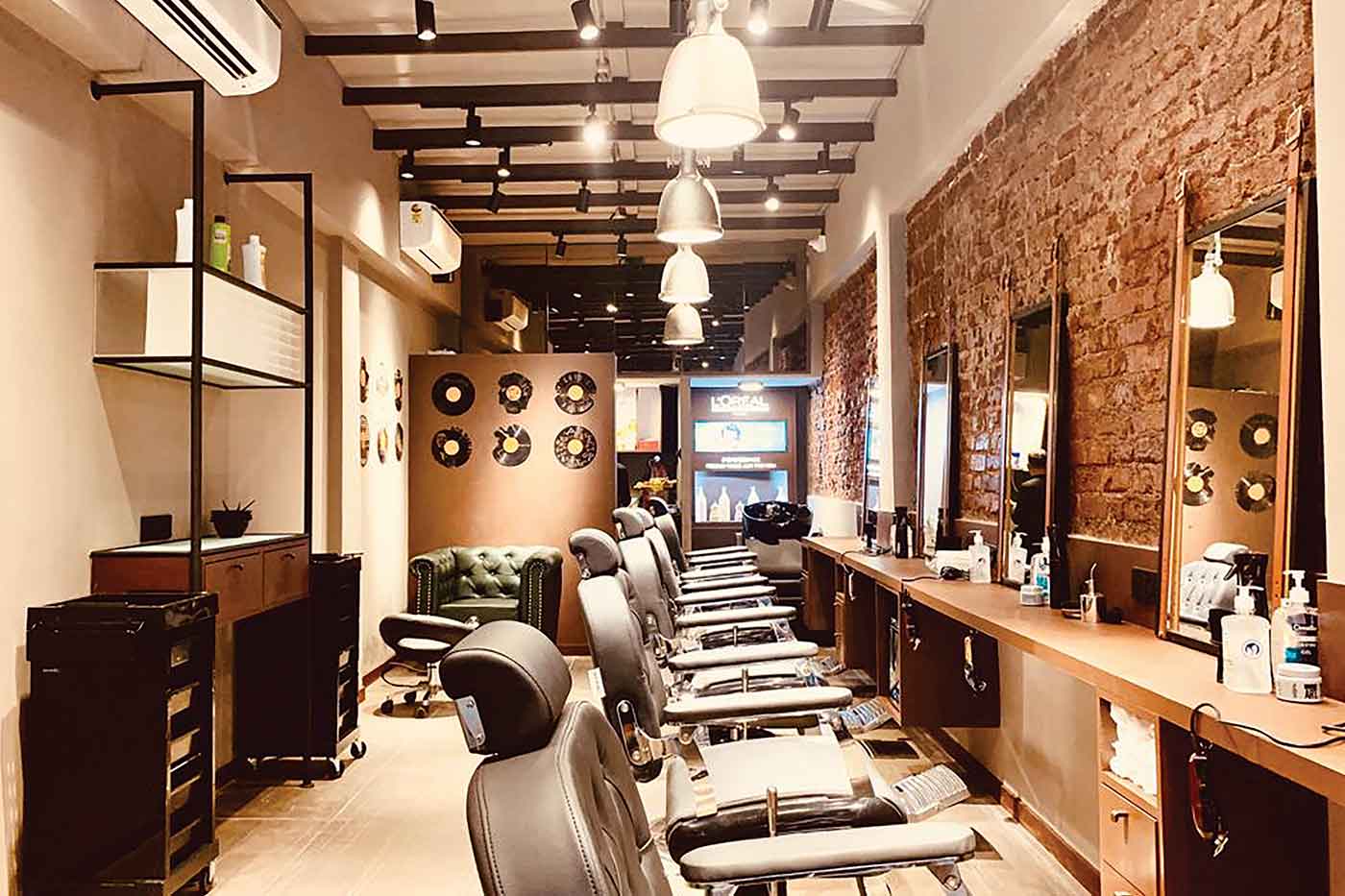 A salon floor with a row of black reclining styling chairs facing a counter and mirrors, an exposed brick wall on the right and pendant lamps overhead