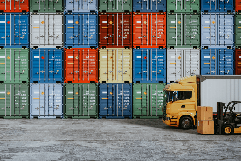 A wall of stacked, multicoloured shipping containers with a yellow truck and a forklift carrying cardboard boxes in front of it