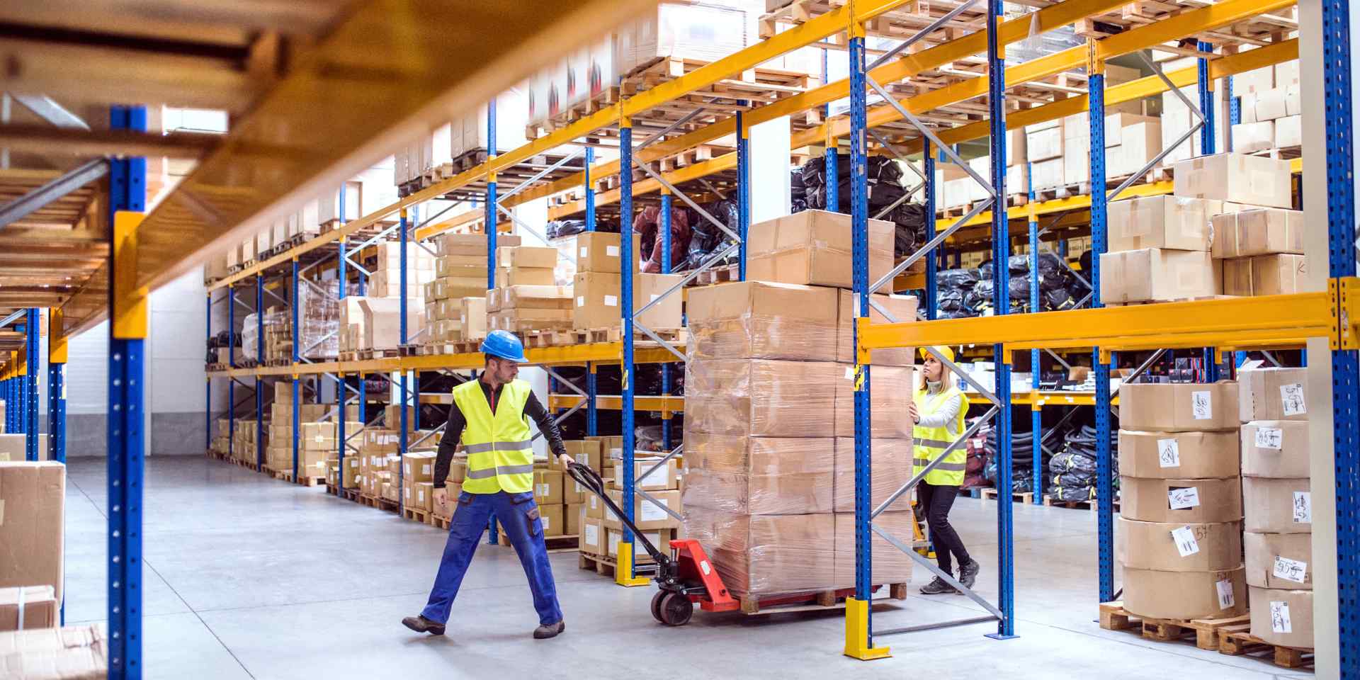 Two warehouse workers in high-visibility vests moving a shrink-wrapped pallet of boxes with a hand pallet jack between tall storage racking