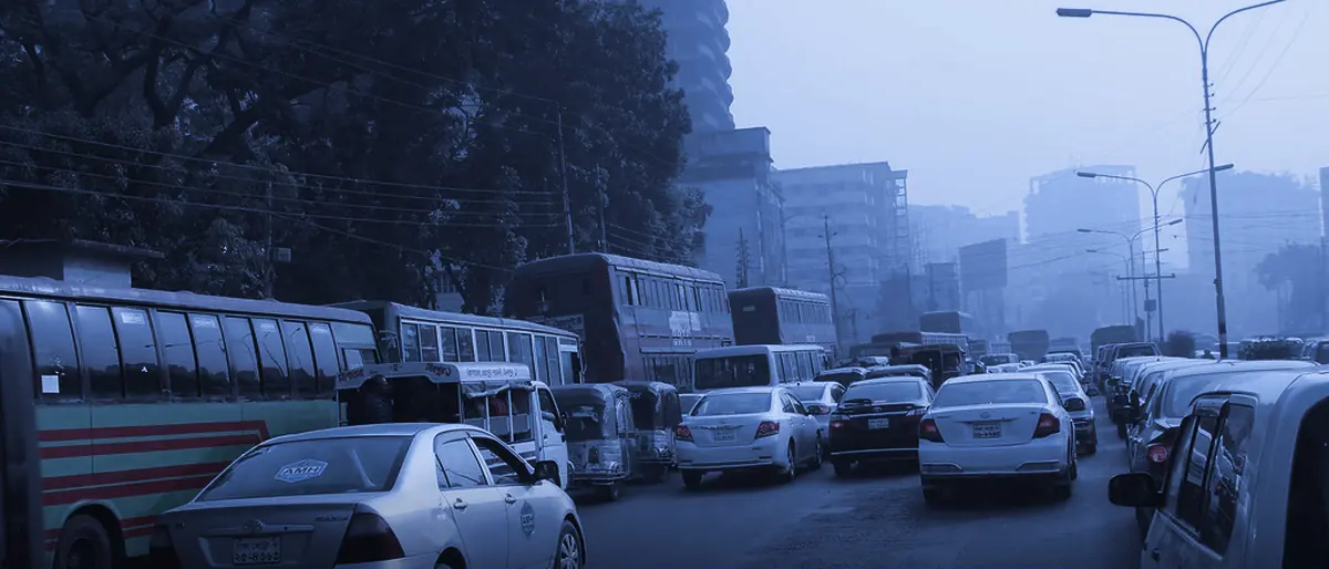 A wide arterial road in Dhaka in daylight, filled with queued buses, cars and taxis under a hazy skyline.